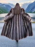 Back view of full-length raccoon fur coat with wide hem, displayed in scenic fjord landscape, premium outerwear for cold seasons.