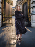 Woman in a fur coat standing in front of an ornate gate on a sunlit street.