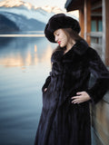 Woman in a black fur coat and hat standing by a body of water with mountains in the background.
