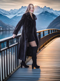 Woman in a long black coat standing on a wooden deck with mountains and lake in the background