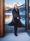 Woman in a long black fur coat standing in front of a large window with a mountain and lake view.