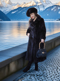 Woman in a long black fur coat standing by a lake with mountains in the background