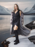 Woman in a long gray coat with fur trim standing by a lake with mountains in the background