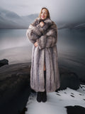 Person wearing a long fur coat standing in a snowy landscape with mountains.