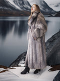 Woman in a fur coat standing in a snowy landscape with mountains and water in the background