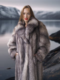 Woman wearing a fur coat standing by a lake with mountains in the background
