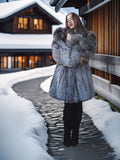 Woman in a fur coat standing on a snowy path in front of a wooden building.