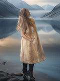 Woman in a fur coat standing by a lake with mountains in the background