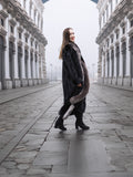 Smiling model in black mink coat with indigo fox trim, walking confidently through a cobblestone street in winter fashion.