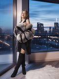 Woman in a fur coat and black boots standing by a large window with a cityscape view at night.