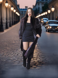 Woman in a black coat and dress walking on a street with vintage cars and streetlights.