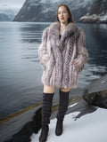 Woman in a fur coat standing by a lake with mountains in the background