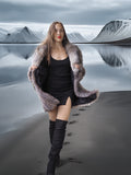 Woman in a fur coat and knee-high boots standing on a reflective surface with mountains in the background