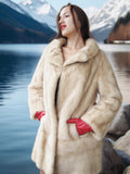 Woman in a fur coat and red gloves standing by a lake with mountains in the background