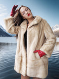 Woman in a fur coat and red gloves standing by a lake with mountains in the background