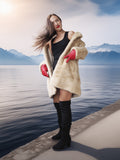 Woman in a fur coat and red gloves standing by a lake with mountains in the background