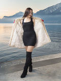 Woman in a black dress and knee-high boots with a fur coat by a lake with mountains in the background