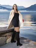 Woman in a fur coat standing by a lake with mountains in the background