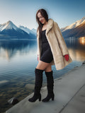 Woman in a fur coat and knee-high boots standing by a lake with mountains in the background