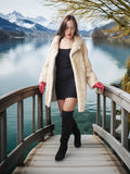 Woman in a fur coat and black dress standing on a wooden bridge over a lake with mountains in the background