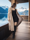 Woman in a long beige coat standing on a wooden deck with mountains and lake in the background