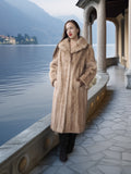 Woman in a fur coat standing by a lakeside with mountains in the background