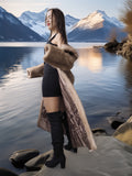 Woman in a fur coat standing by a lake with mountains in the background