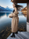 Woman in a fur coat standing by a lake with mountains in the background