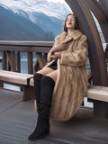 Woman in a fur coat and knee-high boots sitting on a wooden deck with mountains in the background