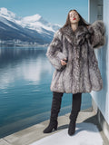 Woman in a fur coat standing by a lake with mountains in the background