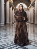 Woman in a long brown fur coat standing in an elegant hallway with columns.