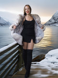 Woman in a fur coat standing by a snowy landscape with mountains and water.