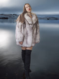 Woman wearing a fur coat standing by a body of water with a dark, overcast sky.