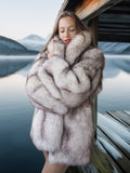 Woman wearing a fur coat standing by a lake with mountains in the background