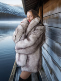 Woman in a fur coat embracing a dog by a wooden cabin by a lake.
