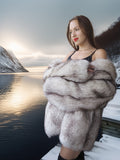 Woman in a fur coat standing by a lake with mountains in the background