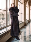 Woman in a long dark fur coat standing in a grand hallway with large windows.