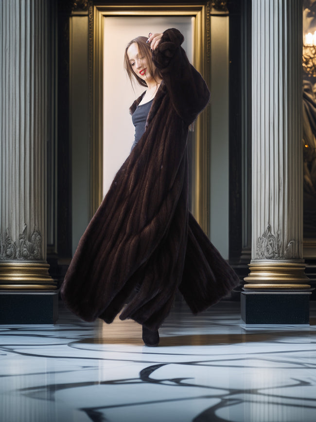Woman in a long brown coat standing in a grand hallway with columns.