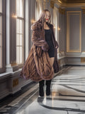 Woman in a long brown fur coat standing in an elegant interior setting.