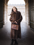 Woman in a long brown fur coat holding a matching handbag in an architectural setting.