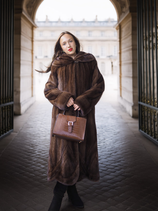 Woman in a long brown fur coat holding a matching handbag in an architectural setting.