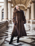 Woman in a long brown fur coat standing in an elegant interior setting.