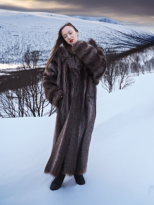 Model in Silver Tip Canadian Raccoon Fur Coat standing in snow-covered hills. Warm and stylish fur outerwear for winter.