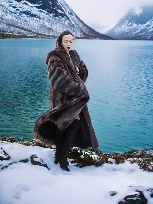 Model in Silver Tip Canadian Raccoon Fur Coat by turquoise lake. Luxurious full-length fur coat for winter elegance.