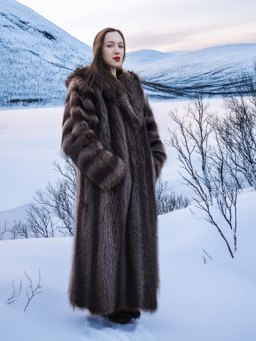Model in full-length Silver Tip Canadian Raccoon Fur Coat standing in snowy valley. Elegant and warm fur coat for winter.