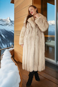Woman in a fur coat standing in front of a wooden cabin with snowy mountains in the background