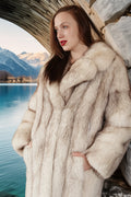 Woman wearing a fur coat standing by a lake with mountains in the background