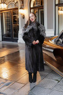 A woman wearing a long black fur coat with a gray and white fur collar stands on a wet stone pavement next to a gold car in front of a building entrance.