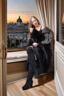 A woman sits on a window seat in a luxurious room, wearing a black faux fur coat with gray fur trim, a black spaghetti strap dress, and black over-the-knee boots, with a sunset view of a historic building in the background.