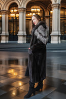 A woman wearing a long dark fur coat with a gray and white fur collar, standing in a grand hall with columns and warm hanging lights.